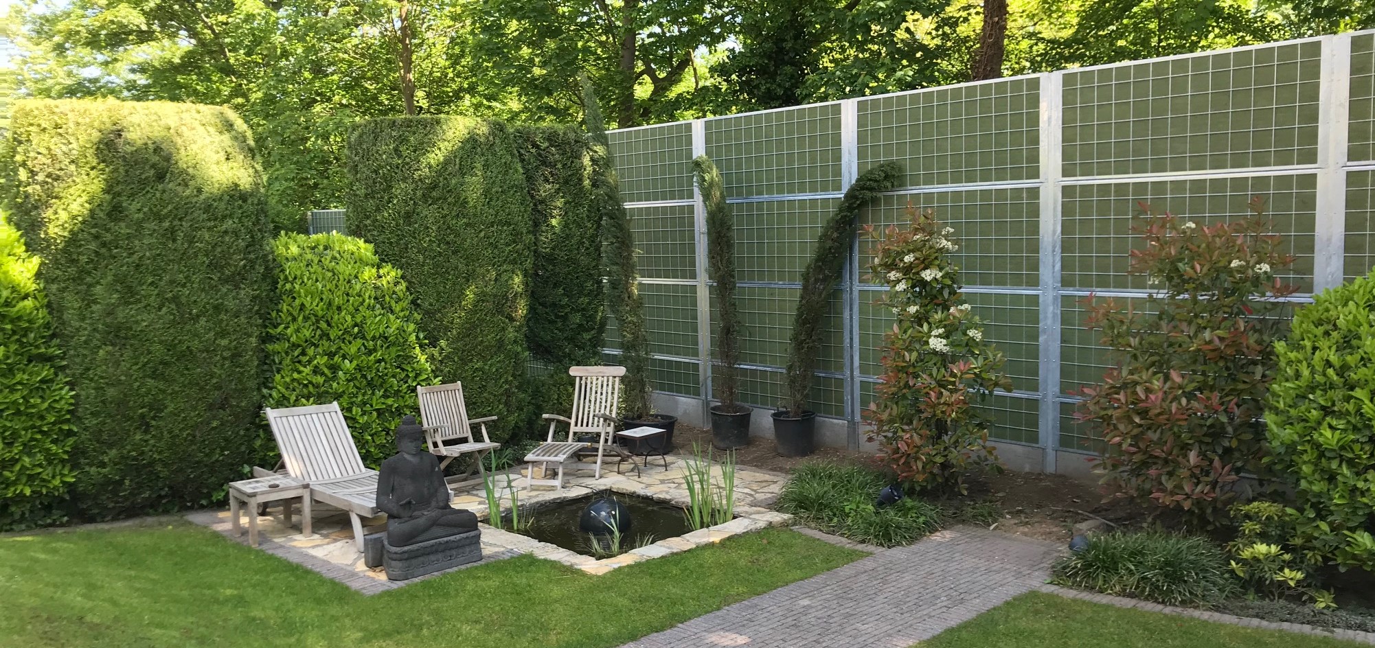 Een gezellige tuin met struiken en een kleine visvijver afgeschermd van burenoverlast met een Noistop® Steel geluidswerende schutting van Poda Omheiningen